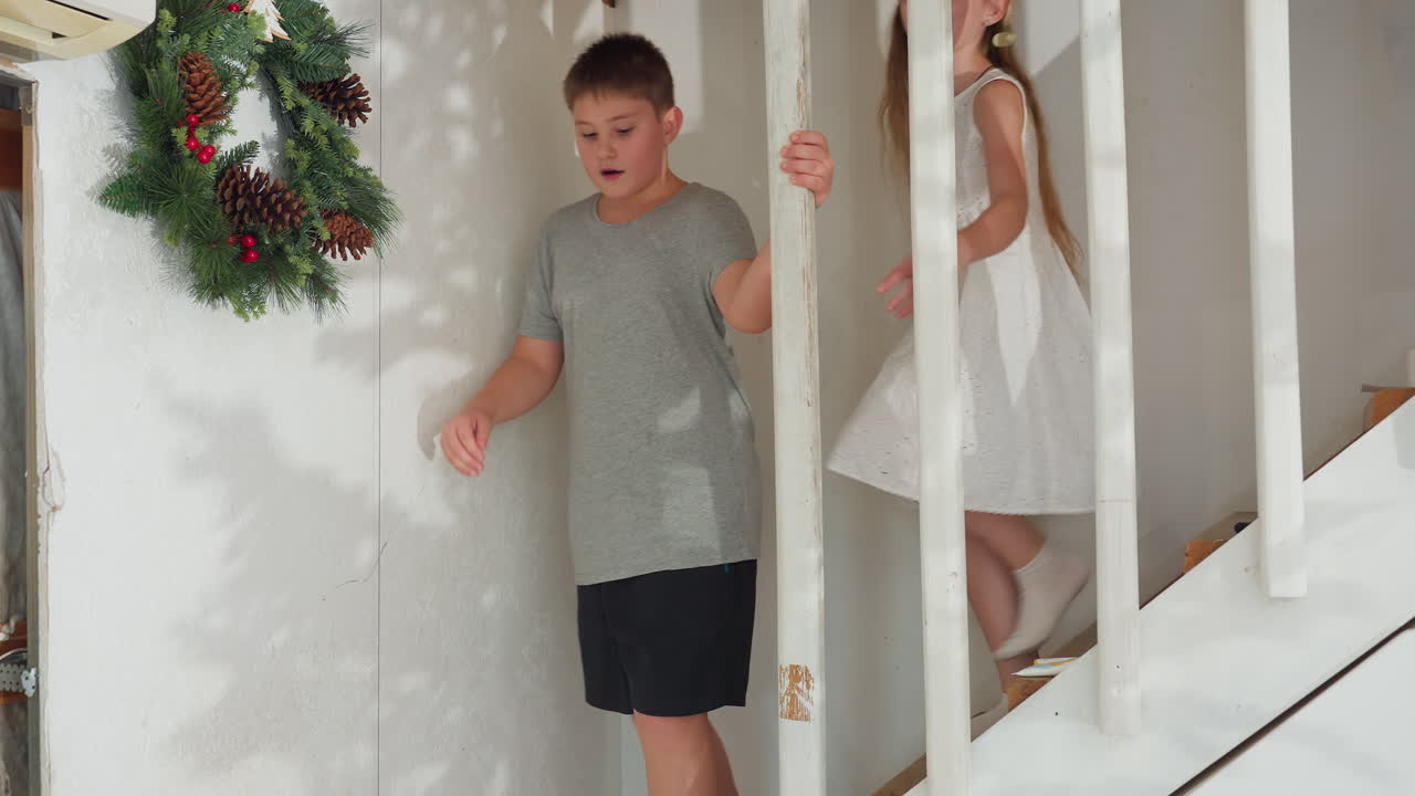 kids in white socks, boy in gray top and black shorts, girl in white gown coming down staircase, boy with surprised expression, girl smiling, playful indoor family moment, festive holiday decorations