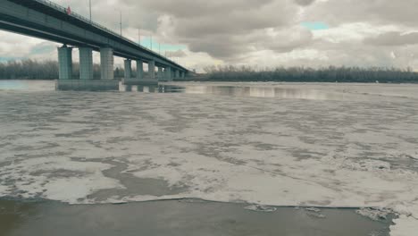 Ancho-Río-Gris-Agua-Cubierta-De-Hielo-Bajo-Un-Enorme-Puente