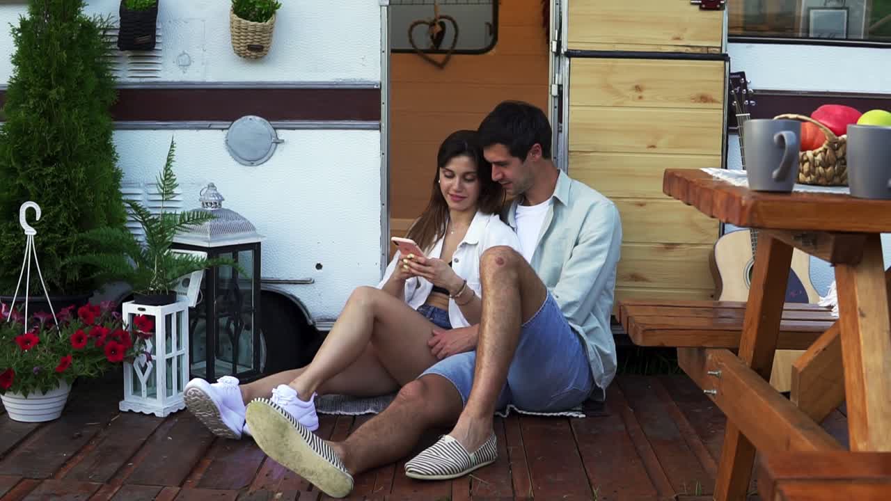 Bella coppia seduta sul pavimento vicino alla porta del camper, che guarda un video sullo smartphone e sorride. Si godono la compagnia, le vacanze condivise, viaggiando in camper.