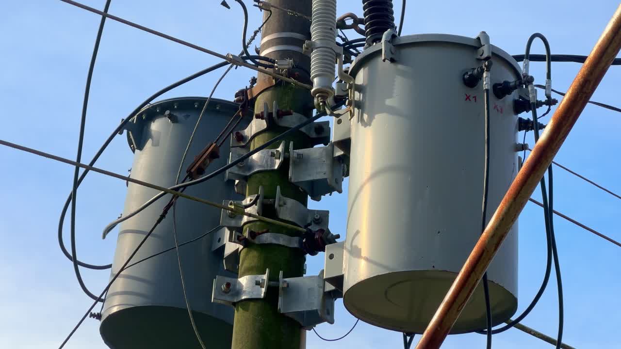 Close-up footage of an electrical transformer mounted on a utility pole, surrounded by tangled high-voltage power lines under a clear blue sky