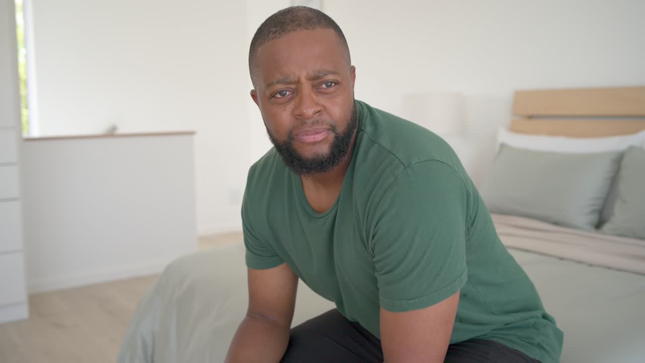 Man in 30s sitting on bed edge in bedroom, listening to off-camera voice and adjusting smartwatch
