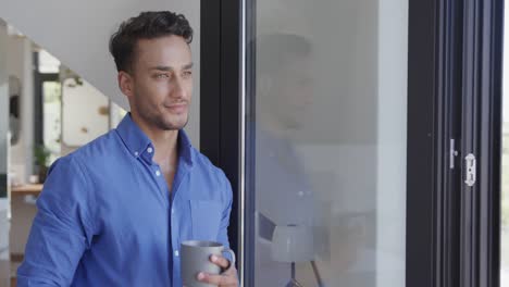 Fröhlicher-Biracial-Mann-Hält-Kaffee-In-Der-Hand-Und-Schaut-Durch-Das-Fenster-In-Einer-Modernen-Wohnung,-Zeitlupe