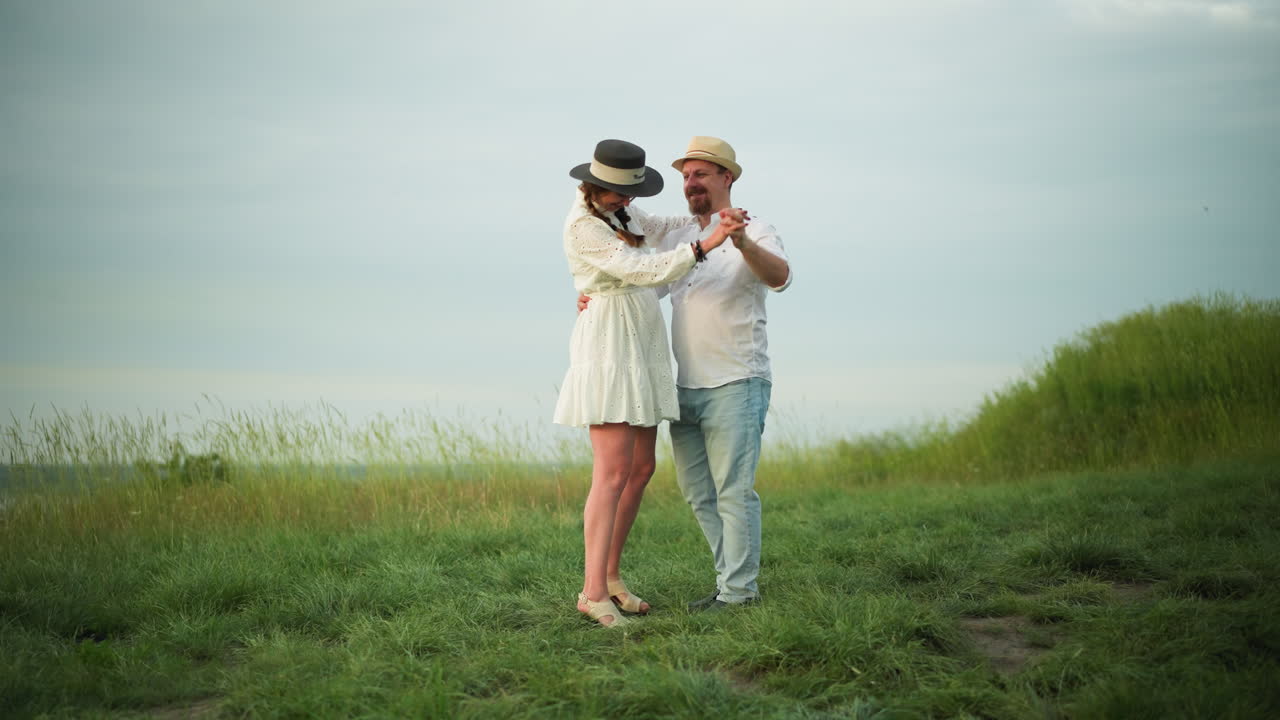 A couple dances together on a grassy hill, with the man wearing a white shirt, hat, and jeans, while the woman is dressed in a white dress and black hat. The serene outdoor setting captures their joy