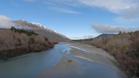 Un-Gran-Río-Fluye-Entre-Un-Bosque-Cerca-De-Queenstown,-Con-La-Notable-Cordillera-En-El-Horizonte