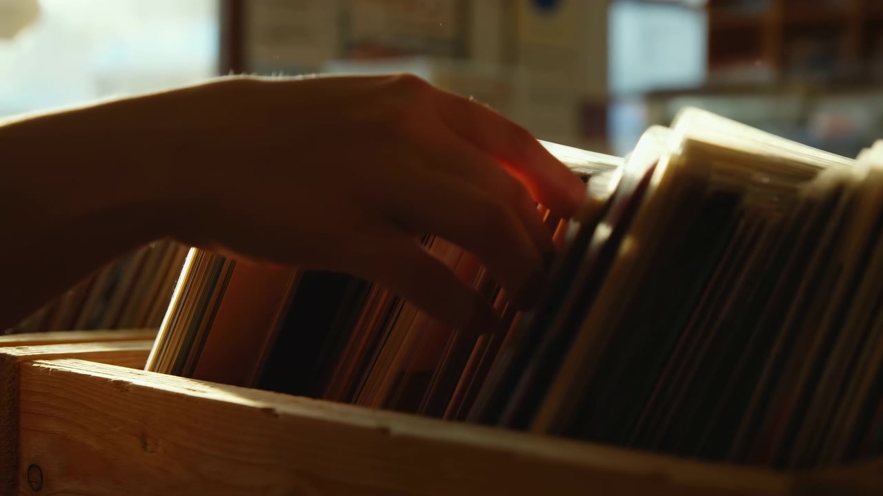 Una mano si protende da sinistra per sfogliare dischi in vinile in verticale conservati in una cassa di legno, con luce solare calda e uno sfondo interno leggermente sfocato che crea un'atmosfera tranquilla
