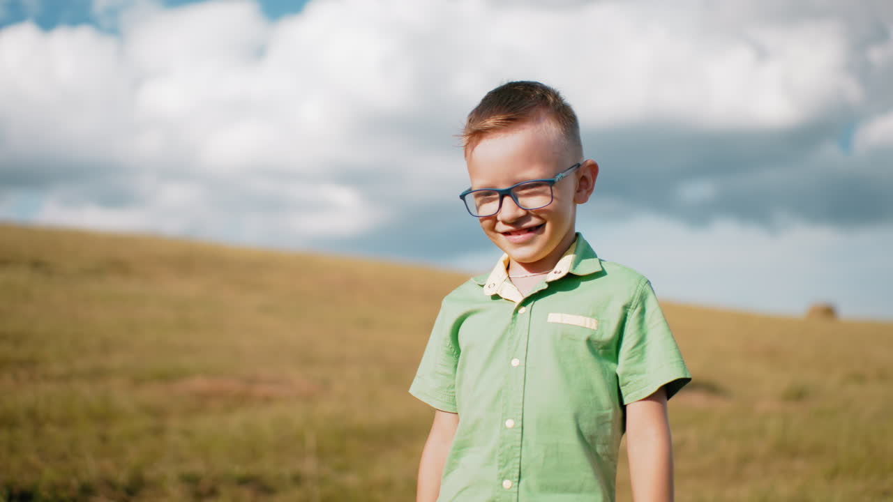 안경과 초록색 셔츠를 입은 작은 소년은 빛이 그의 얼굴에 반사되는 동안 기게 미소 짓고, 그는 열린 하늘과 따뜻한 시골 분위기로 둘러싸인 풀으로 된 광활한 농장에서 서 있습니다.