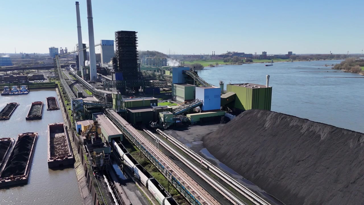 Un drone avanza verso un grande cumulo di carbone e un impianto industriale costruito su una penisola fluviale.
