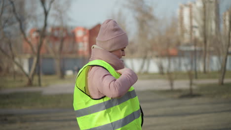 Woman in pink winter jacket and knit beanie standing outdoors in city park, fastening bright yellow reflective safety vest over warm clothing on sunny cold day, preparing for outdoor work activity