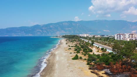 aerial drone panning low across a highway towards a white sandy tropical beach with the mediterranean sea and a coastal beach town in finike turkey on a sunny summer day