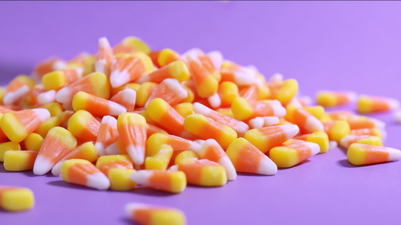 Classic sweet candy corn pieces for Halloween falling in slow motion onto a pile against a vibrant purple background, creating a festive and colorful scene perfect for seasonal treats