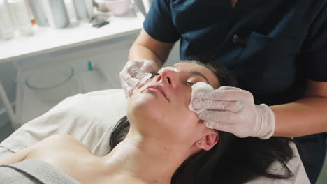 Close up view of wellness technician using cotton pads to clean visitor face while visitor closes eyes peacefully during facial treatment in bright spa room with soft decor and ambient lighting