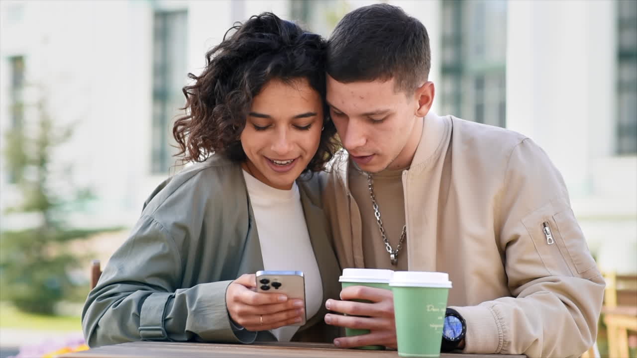 A happy couple outdoors near a cafe. Looking in the phone, smiling, talking and kissing, coffee. Autumn atmosphere. Slow motion