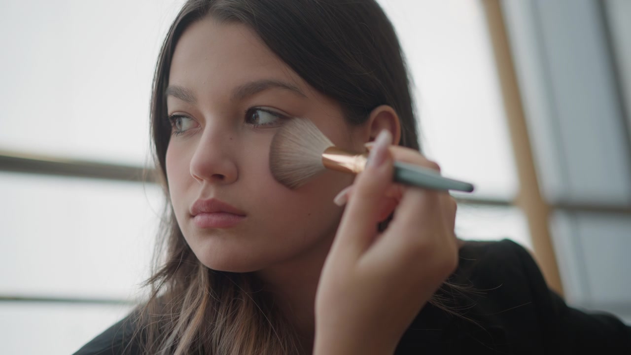 Close low-angle portrait shows young woman applying makeup with large brush, hand moving across cheek and jawline while blending powder, creating polished beauty routine in soft indoor light