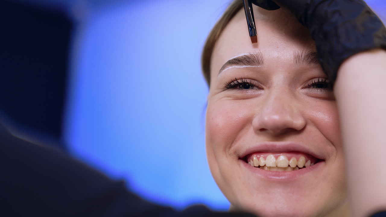 Happy smiling Caucasian girl in beauty salon having beauty procedure. Cosmetologist uses a brush to make straight lines around girl's eyebrows. Close up.