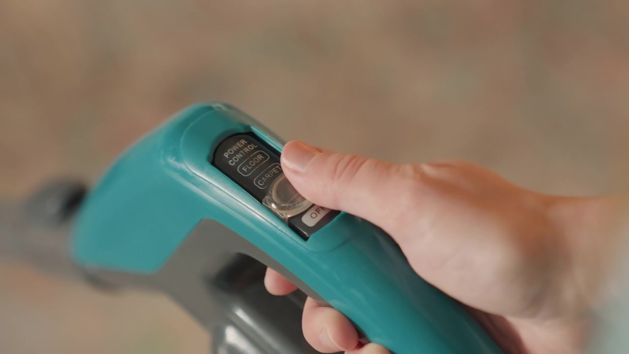 Closeup hands adjusting vacuum dial, turquoise cordless vacuum handle, thumb turning power setting, indoor carpet background, homeowner preparing house cleaning routine, soft natural light, modern