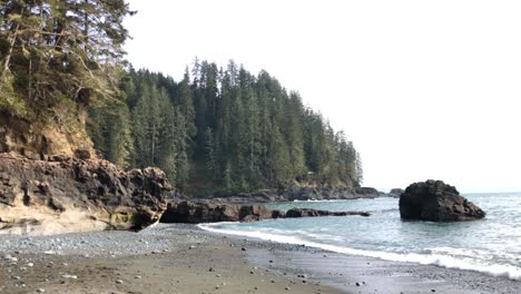 Sea-waves-hitting-rugged-sand-shore-surrounded-by-dense-pine-tree-forest-on-an-overcast-day,-Sombrio-Beach,-British-Columbia,-Canada