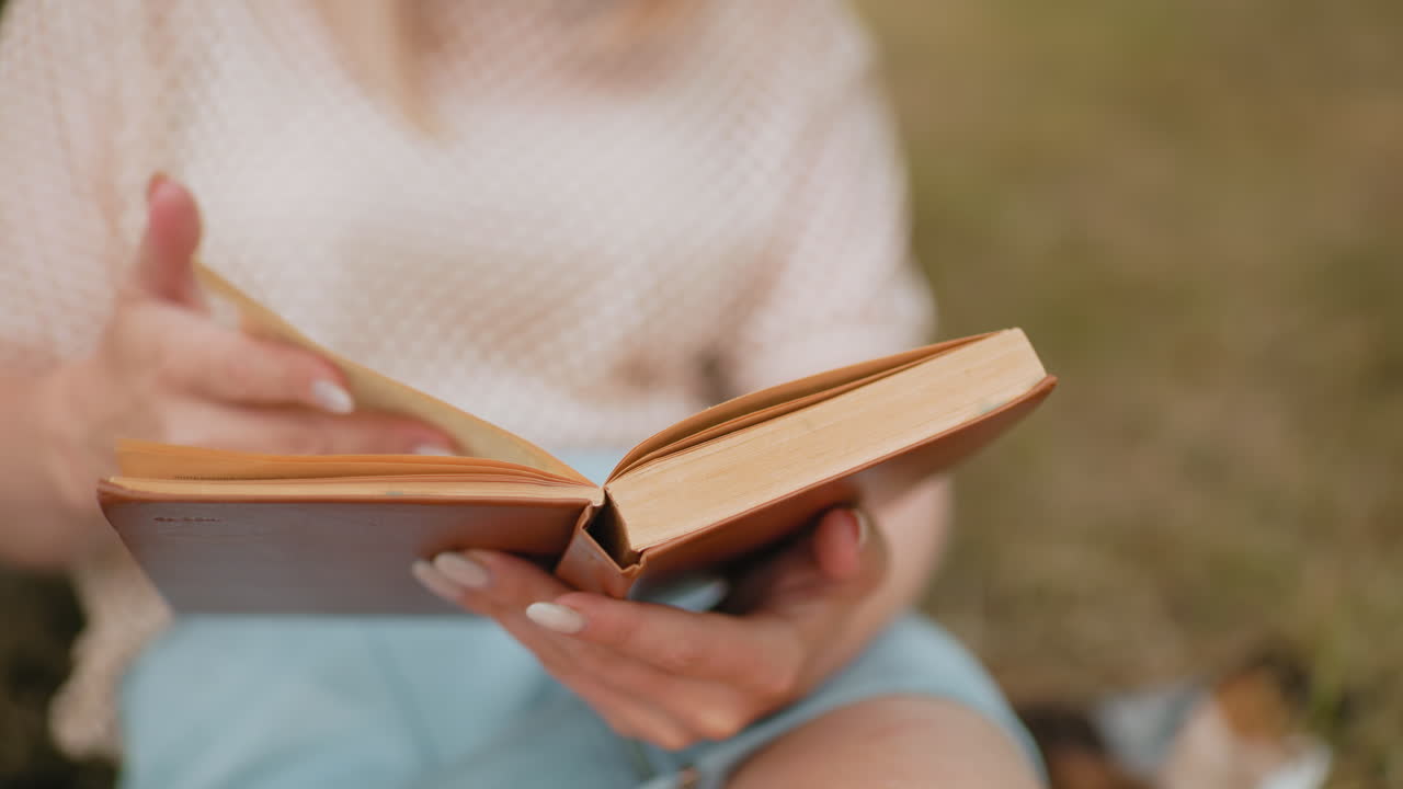 primer plano de manos volviendo páginas en un libro abierto, enfoque suave en pantalones cortos azules y uñas delicadas momento de lectura al aire libre pacífico con un fondo natural borroso