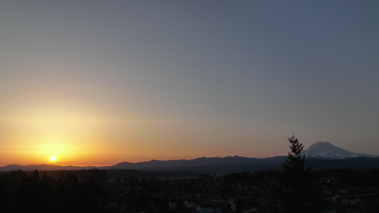 Ascending aerial revealing the sun rising over Mount Rainier with trees in the foreground.