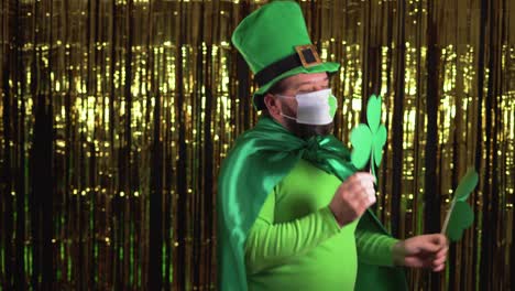 a young leprechaun celebrates st patrick's day during the pandemic by wearing a medical mask. dancing with clover leaves in her hands.