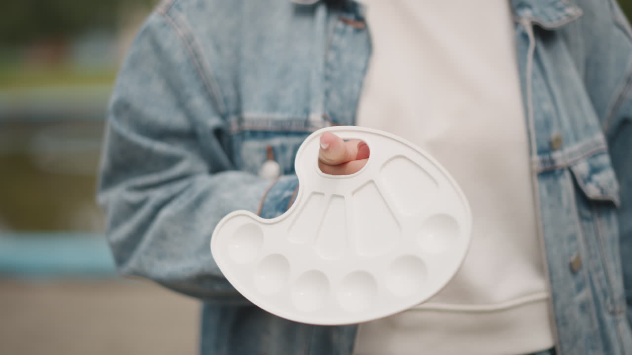 Closeup artist holding paint palette outdoors denim jacket white hoodie finger through palette hole empty white mixing wells soft daylight park railing background calm preparation for painting casual