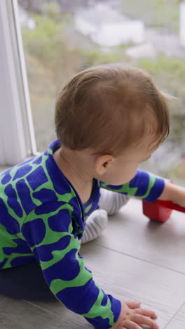 little adorable baby boy playing with a red dumb-bell on the floor. lovely toddler crawling by the floor at big panoramic window. high angle view. vertical video