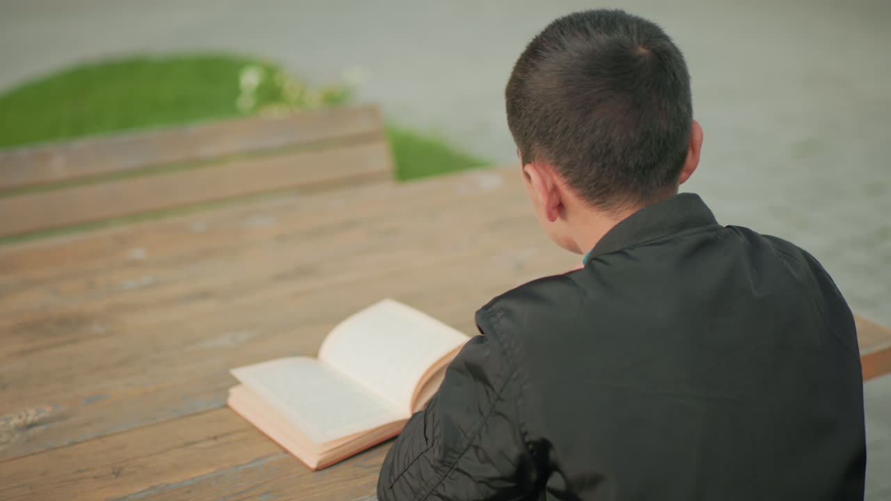 Side view of boy seated at wooden table outdoors writing in notebook with open book in front of him, while blurred bicycle passes by in background, symbolizing focus amid movement