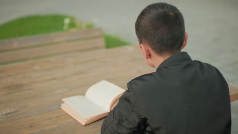side view of boy seated at wooden table outdoors writing in notebook with open book in front of him, while blurred bicycle passes by in background, symbolizing focus amid movement