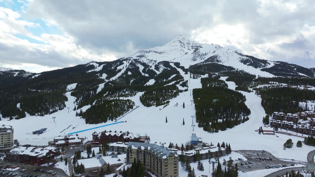 Plano aéreo amplo de drone em ascensão de uma paisagem de inverno de um resort de montanha em Montana, com o Lone Peak elevando-se ao fundo, cercado por florestas de pinheiros, casas e terreno coberto de neve.