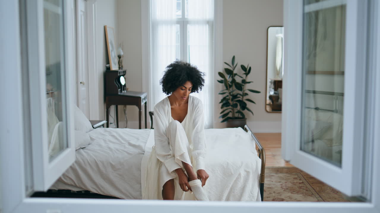 Ragazza serena che indossa calzini nella camera da letto al mattino. Signora riccia spensierata, routine quotidiana.