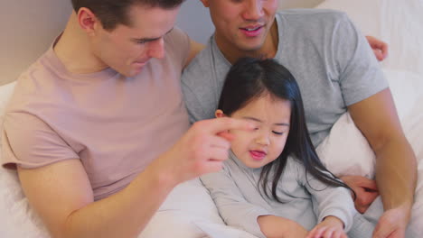 Familia-Con-Dos-Padres-En-La-Cama-En-Casa-Leyendo-Un-Cuento-A-Su-Hija