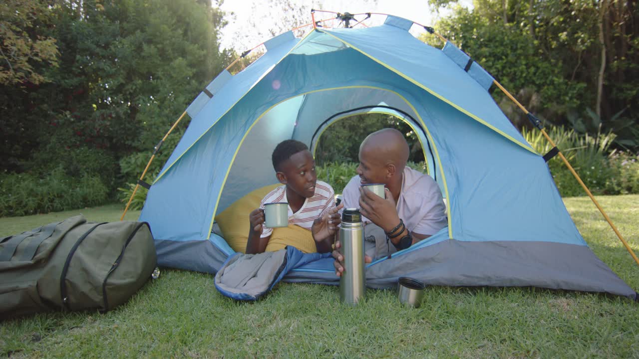 Afrikansk far og søn slapper af med krus, mens faren sætter en termokande, og sønnen nipper til sin drik i et blåt telt.