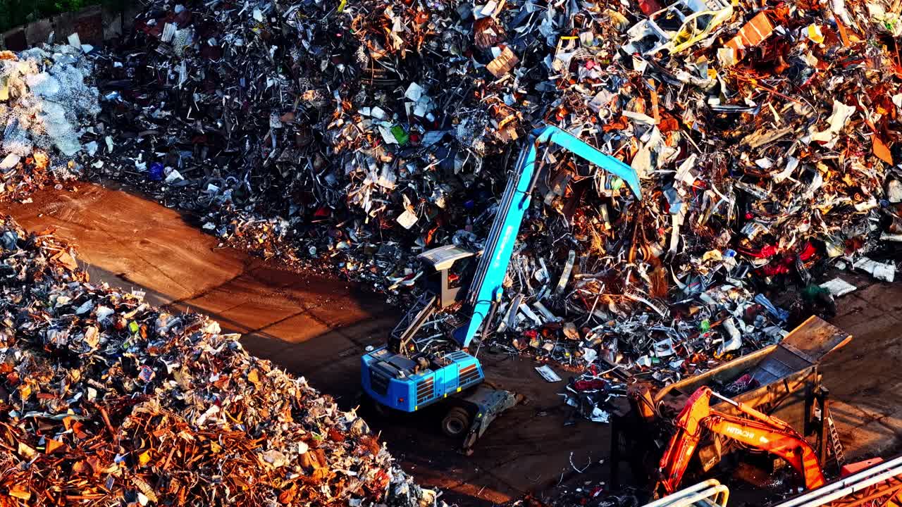 Vista aerea di un operatore di materiali blu che seleziona un enorme cumulo di rottami metallici misti in un deposito industriale di riciclaggio, circondato da mucchi di detriti frantumati e macchinari