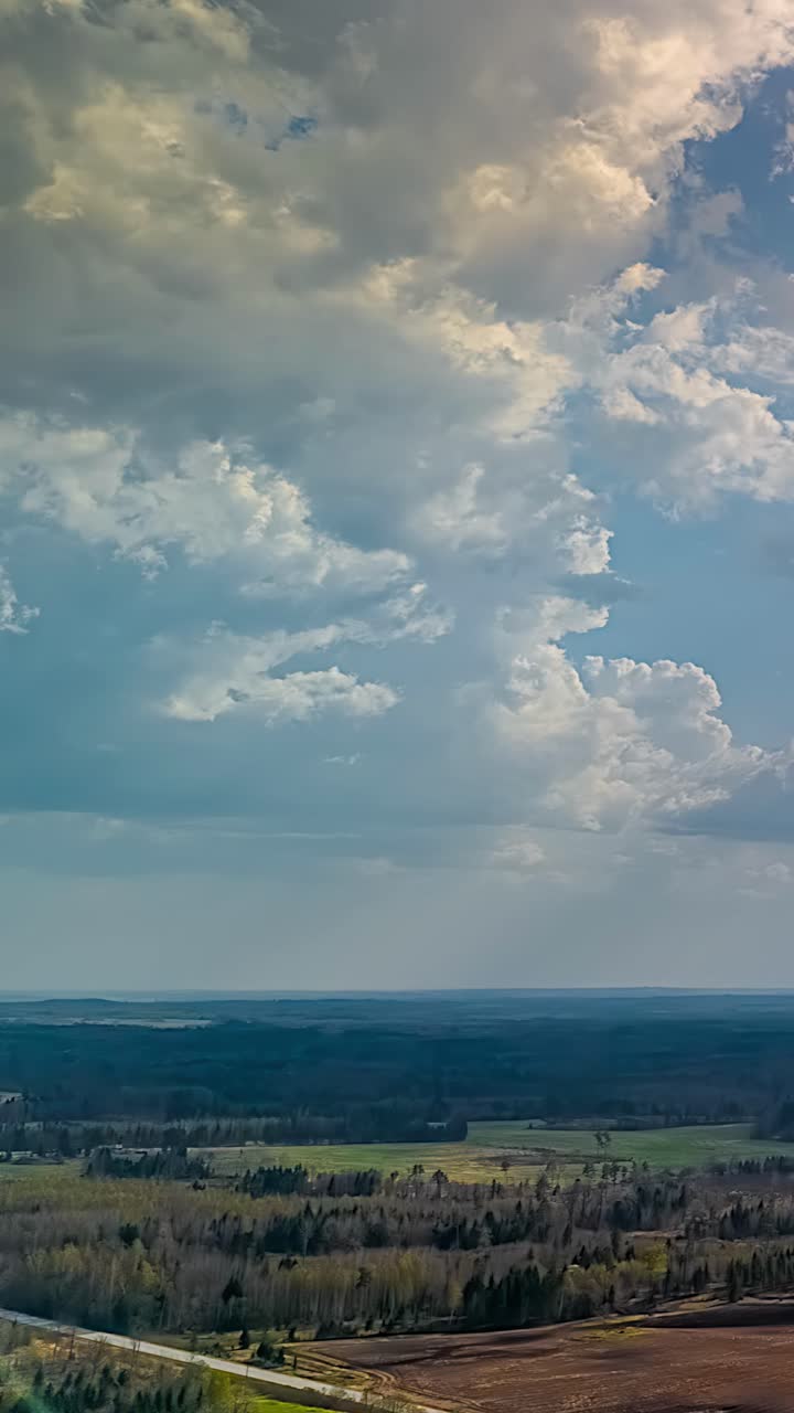 A dynamic vertical aerodynamic timelapse showing white cumulus clouds moving quickly over endless green meadows and forest landscape under a blue sky