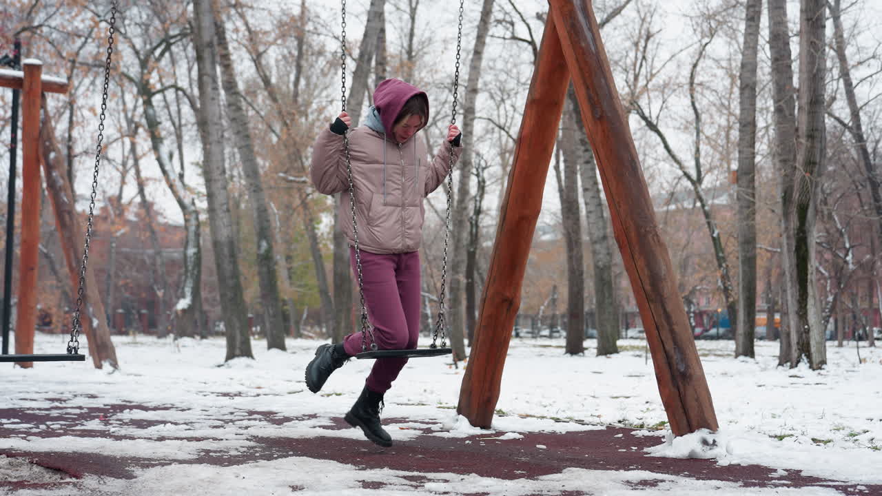 joven vestida de invierno coloca cuidadosamente la rodilla en un columpio de hierro mientras sostiene cadenas para el equilibrio, suelo cubierto de nieve, árboles desnudos y edificios residenciales en el fondo