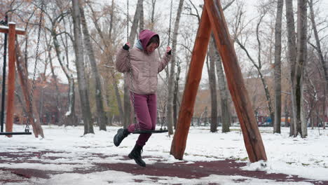 young woman in winter attire carefully places knee on iron swing while holding chains for balance, snow-covered ground, bare trees, and residential buildings in background