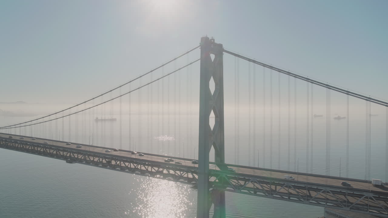 Traffic crosses suspension bridge over water as mist obscures boats on the horizon