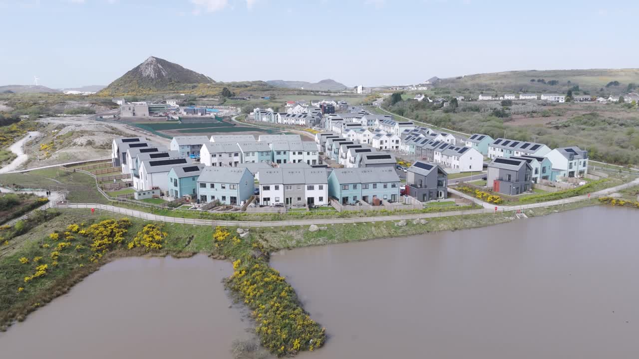 Aerial sweep of eco housing estate with pastel facades, solar roofs and a retention pond set against a reclaimed quarry mound for sustainable waterfront living