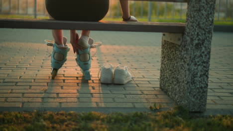 back view of lady seated on bench in warm evening light removing right roller skate with one hand resting on bench, two white sneakers on ground beside her, sunset glow, and blurred iron railing