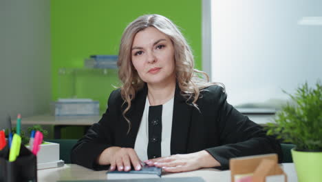 business expert seated at modern office desk with organized workspace including laptop, stationery, architectural model, and potted plant, looking directly at camera with composed expression