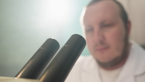 a close-up view of a technician using a microscope in a laboratory, the foreground shows the microscope lenses as he placed his eyes on the microscope, with light on behind him