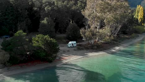 Motorhome-on-beach-beautiful-blue-Lake-Wakatipu,-Queenstown,-New-Zealand-and-trees-scenery-mountains-with-fresh-snow-during-fall-autumn---Aerial-Drone