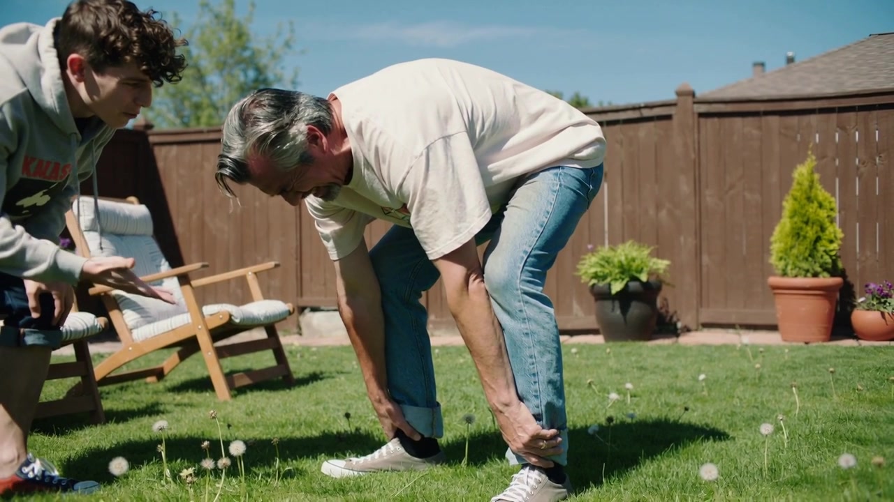 Bending senior man feeling discomfort adjusting jeans and sneakers on lawn by fence teen gesturing