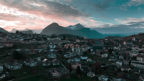 Breathtaking-drone-flight-over-Spiez,-Switzerland-at-sunset,-showcasing-mountains,-houses,-green-hills,-trees,-and-a-stunning-sky