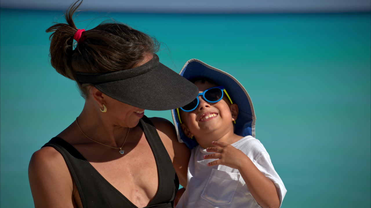 Portrait of a mexican latin brunette woman wearing a visor and sunglasses carrying her son looking at the camera smilling at the beach in Cancun