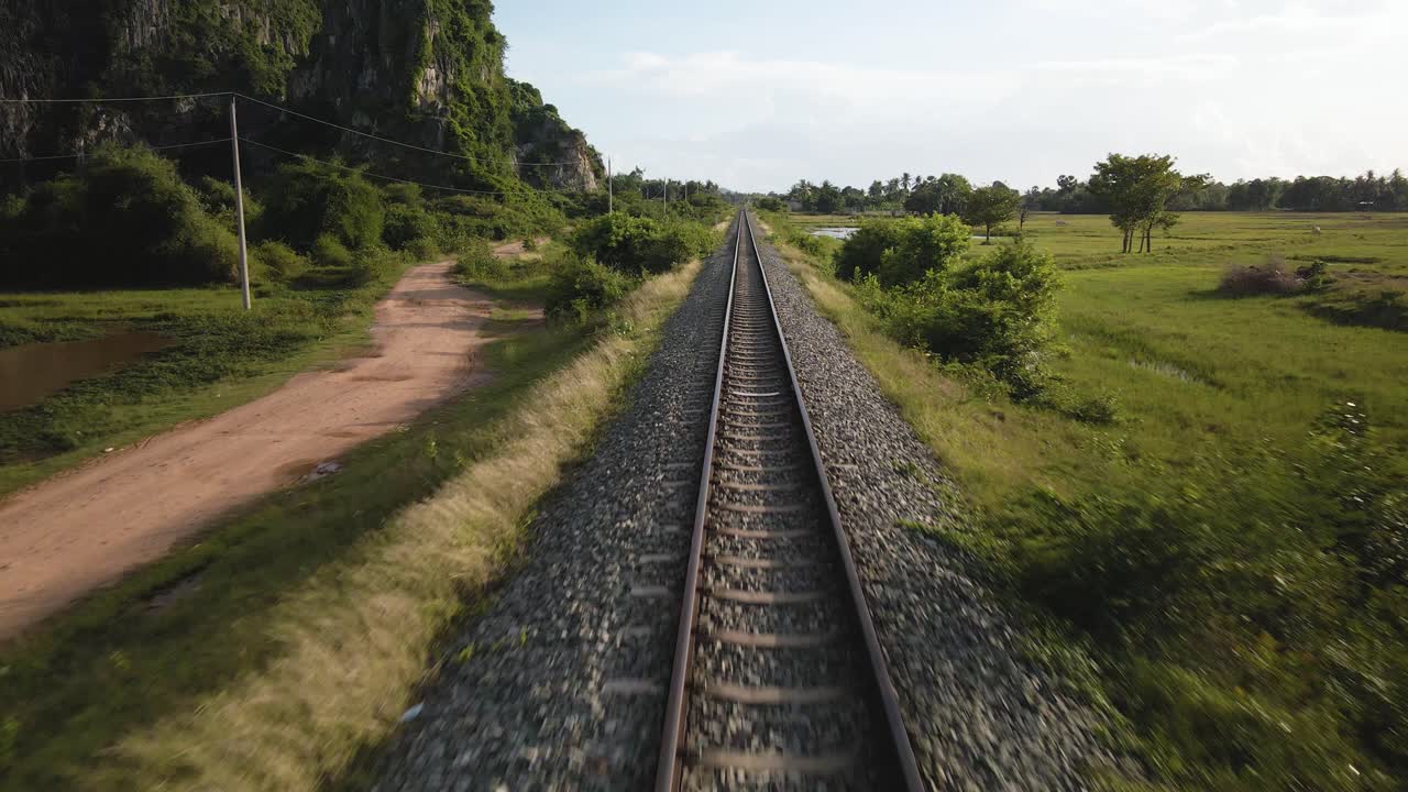 Jalur kereta api rel sempit lurus membentang menuju perspektif satu titik di cakrawala, drone terbang di atas pedesaan Kamboja, Asia Tenggara