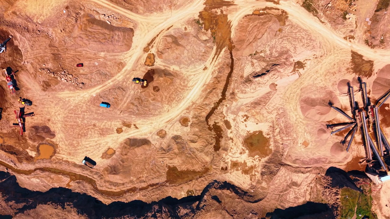 Top down aerial view of sand processing plant and conveyor belts. Sorting machinery and conveyor systems stand near sand piles and vehicle tracks on brown ground during bright daylight.