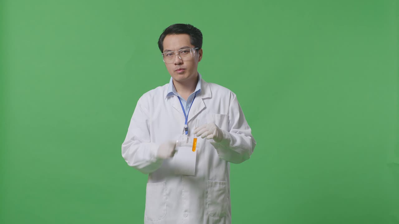 Asian Man Scientist With Orange Liquid In The Test Tube Looking At Camera And Making Shh Gesture While Standing On The Green Screen Background In The Laboratory
