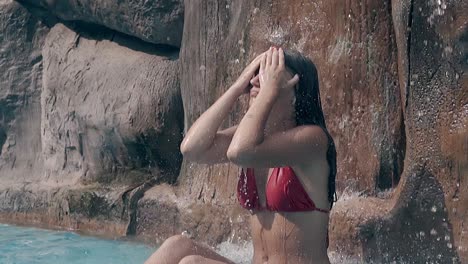 dama en bikini rojo se sienta debajo de una cascada artificial en la piscina