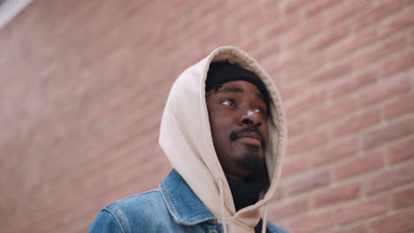 handsome young man wearing hoodie layered under denim jacket walking along red brick wall outdoors on urban sidewalk looking upward thoughtfully