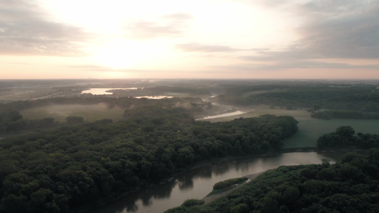 Cinematic drone flight follows a calm river surrounded by trees and natural Midwestern scenery.
